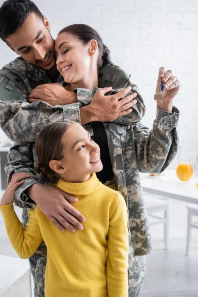 Niño sonriente mirando la llave cerca de los padres en uniforme militar abrazándose en casa - foto de stock