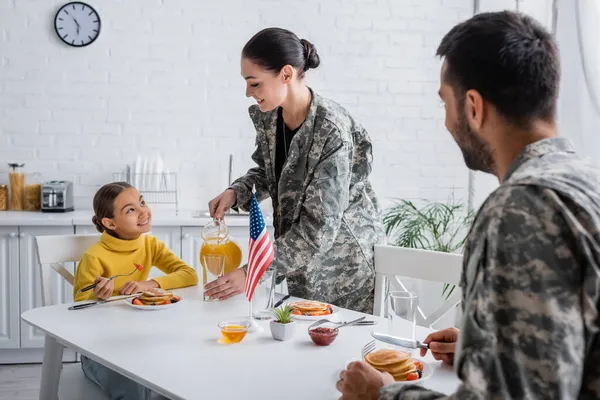 Mujer sonriente en uniforme militar vertiendo jugo de naranja cerca de la familia y la bandera americana en la mesa en casa - foto de stock