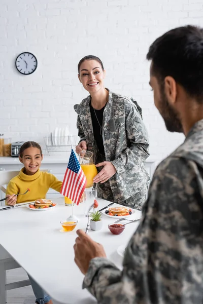 Smiling woman in military uniform holding orange juice near daughter and husband during breakfast at home — Stock Photo