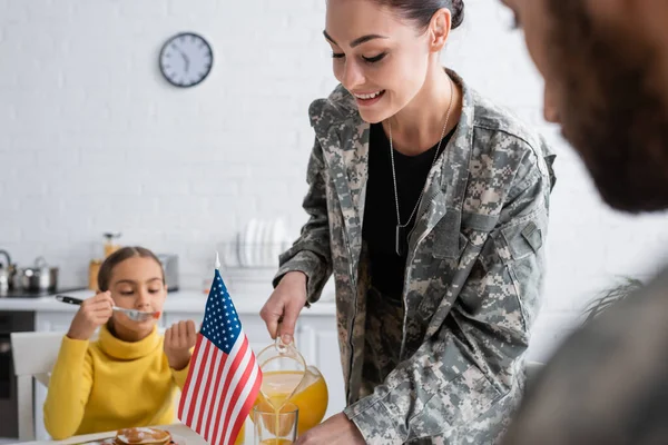 Lächelnde Frau in Militäruniform gießt Orangensaft neben verschwommener Familie und amerikanischer Flagge zu Hause aus — Stockfoto