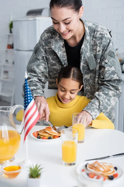 Smiling woman in camouflage uniform cutting pancakes near daughter and american flag in kitchen — Stock Photo