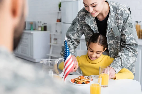 Smiling mother in camouflage cutting pancakes near daughter and blurred husband at home — Stock Photo