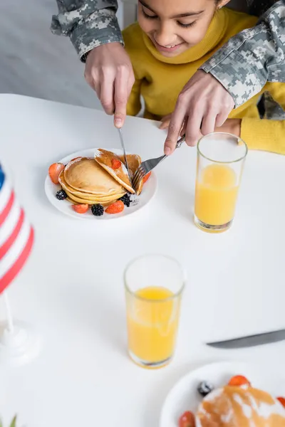 Mère en camouflage militaire coupant des crêpes près de fille souriante et drapeau américain à la maison — Photo de stock