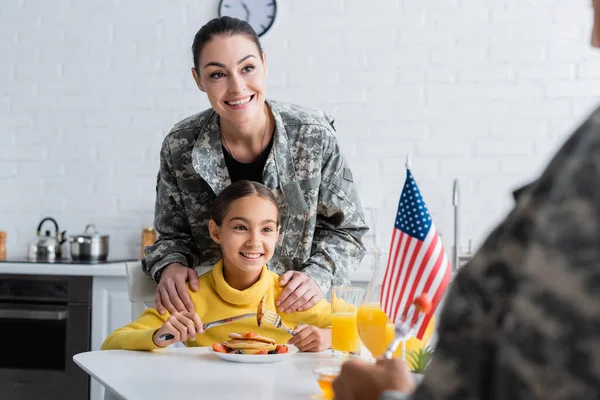 Smiling mother in camouflage uniform standing near daughter and american flag during breakfast in kitchen — Stock Photo