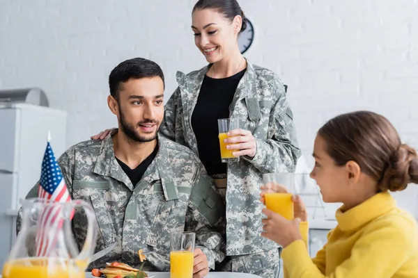 Sonrientes padres en uniforme militar mirando a su hija cerca de la bandera americana durante el desayuno en casa - foto de stock