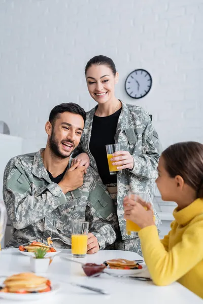 Cheerful parents in military uniform holding hands near daughter during breakfast at home — Stock Photo