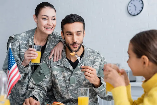 Parents souriants en uniforme militaire regardant un enfant près du drapeau américain pendant le petit déjeuner à la maison — Photo de stock