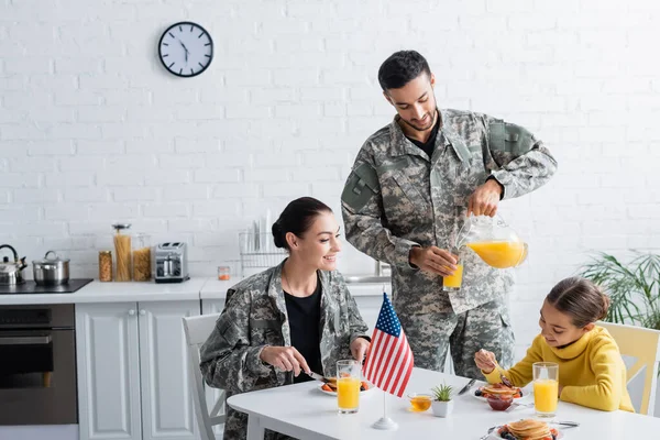Hombre en uniforme militar vertiendo jugo de naranja cerca de la familia y la bandera americana en la cocina - foto de stock