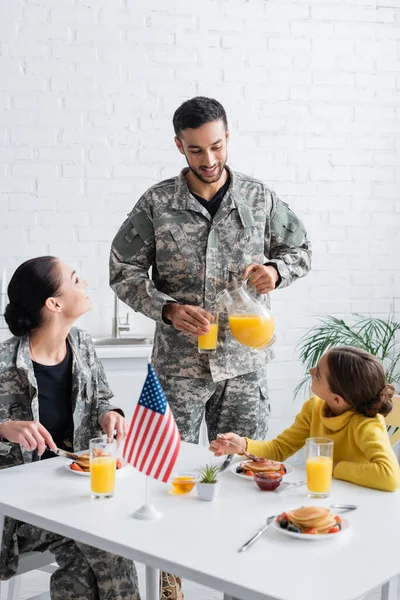Man in military uniform pouring orange juice near family and american flag during breakfast at home — Stock Photo