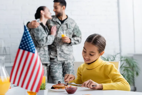 Niño sonriente sentado cerca de panqueques, bandera americana borrosa y padres en uniforme militar en casa - foto de stock