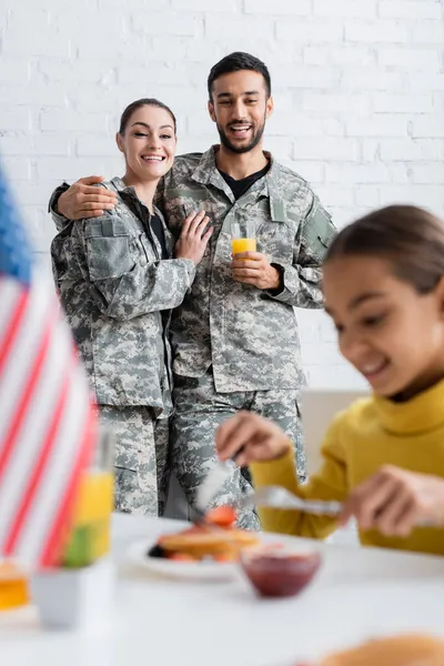 Cheerful parents in camouflage uniform looking at daughter having breakfast near blurred american flag at home — Stock Photo
