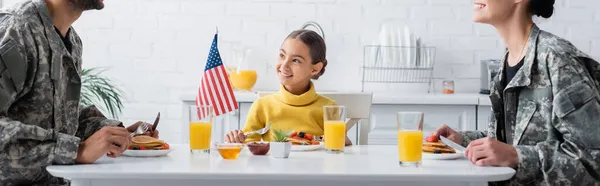 Smiling girl sitting near breakfast, american flag and parents in camouflage, banner — Stock Photo