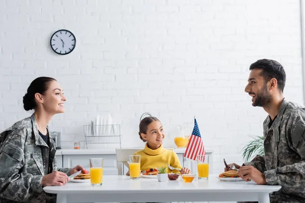 Genitori allegri in camuffamento seduti vicino alla colazione, bandiera americana e figlia a casa — Foto stock