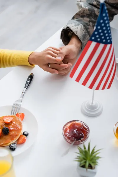 Vista recortada del hombre en uniforme militar cogido de la mano de la hija cerca del desayuno y la bandera americana - foto de stock