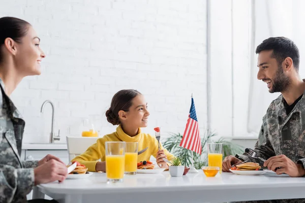 Ragazzo positivo guardando i genitori in uniforme militare vicino gustosa colazione e bandiera americana a casa — Foto stock