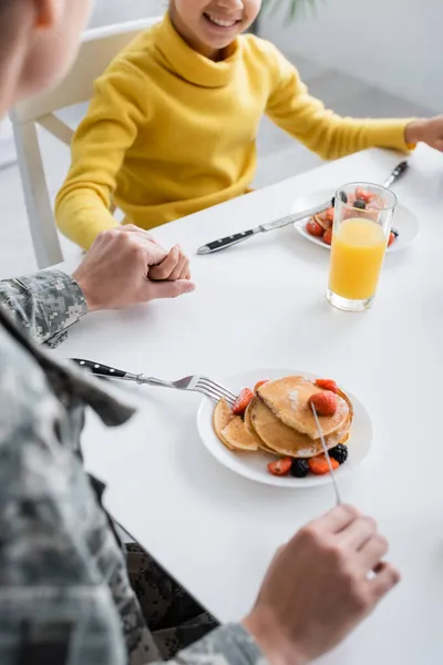 Vista recortada de la mujer en uniforme militar cogida de la mano de un niño sonriente cerca de panqueques y jugo de naranja en casa - foto de stock