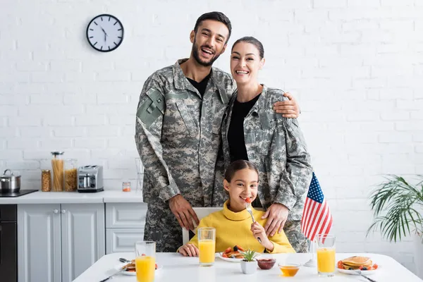 Parents in military uniform looking at camera near child, breakfast and american flag in kitchen — Stock Photo