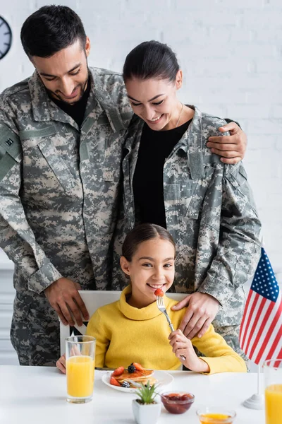 Happy parents in military uniform standing near daughter with breakfast and american flag in kitchen — Stock Photo