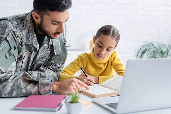 Father in military uniform pointing at book near daughter and laptop at home — Stock Photo