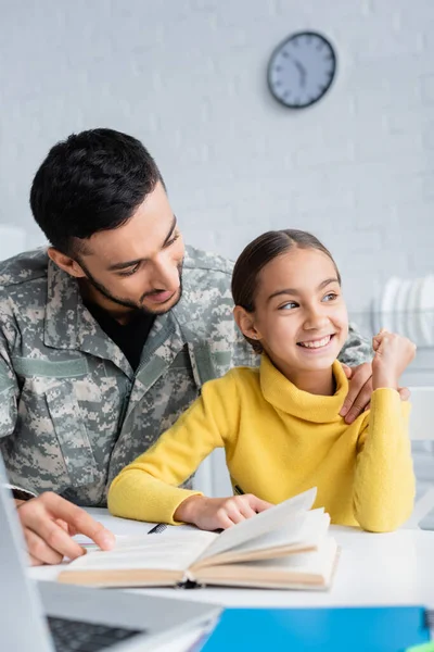 Uomo sorridente in uniforme camuffamento abbracciando figlia vicino libro e laptop a casa — Foto stock