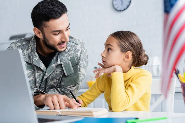 Lächelnder Vater in Tarnuniform zeigt auf Buch neben Tochter, Laptop und amerikanischer Flagge zu Hause — Stockfoto