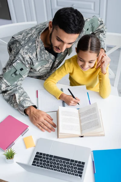 Vista aérea del hombre en uniforme militar sentado cerca de la hija escribiendo en el cuaderno y el ordenador portátil en casa - foto de stock