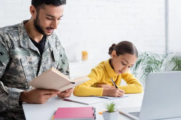 Pai em uniforme militar segurando livro perto da filha escrevendo no notebook e laptop em casa — Fotografia de Stock