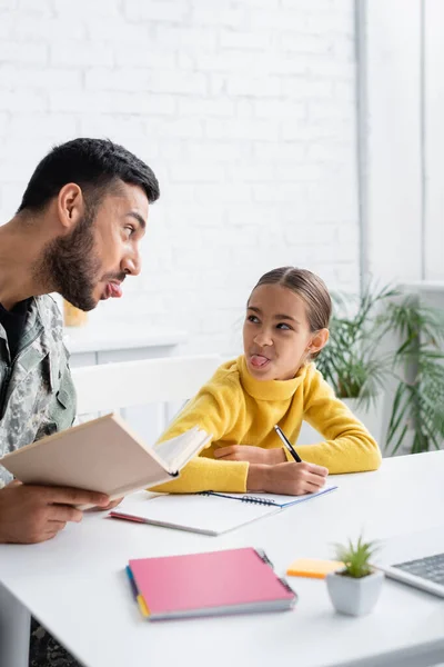 Homme en uniforme militaire tenant le livre près de la fille sortant la langue et écrivant sur le carnet à la maison — Photo de stock