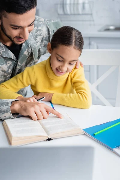 Man in camouflage uniform pointing at book near smiling daughter and laptop at home — Stock Photo