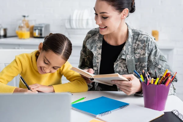 Lächelnde Mutter in Militäruniform hält Buch neben Tochter mit Stift und Notizbuch zu Hause — Stockfoto