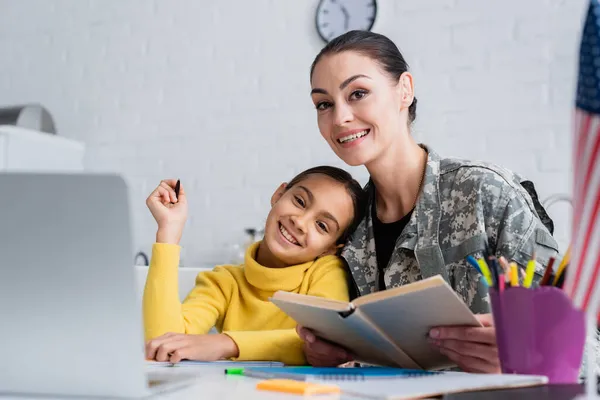 Femme souriante en uniforme militaire tenant le livre près de l'enfant, ordinateur portable et drapeau américain à la maison — Photo de stock