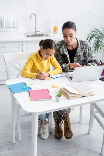 Mother in military uniform looking at laptop while daughter writing on notebook at home — Stock Photo