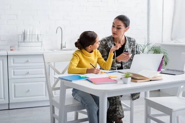 Chica preadolescente escribiendo en un cuaderno cerca de la madre en uniforme militar apuntando a la computadora portátil en casa - foto de stock