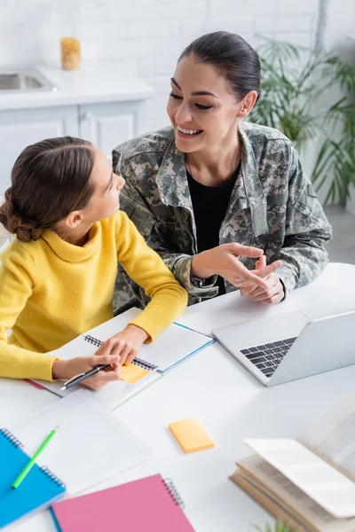 Cheerful woman in military uniform pointing at laptop near daughter and notebooks and home — Stock Photo