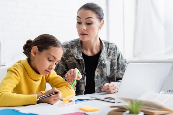 Madre in uniforme militare che indica notebook vicino a figlia e notebook a casa — Foto stock