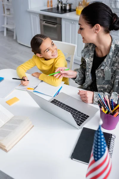 Mãe sorridente em uniforme militar segurando lápis perto da filha com notebook e laptop em casa — Fotografia de Stock