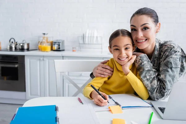 Mujer sonriente en uniforme militar abrazando a un niño cerca de los cuadernos y el portátil en casa — Stock Photo