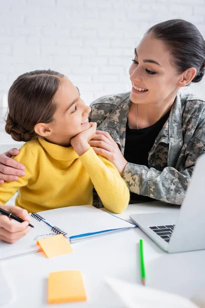 Mère souriante en uniforme militaire regardant l'enfant pendant les travaux scolaires près d'un ordinateur portable à la maison — Photo de stock