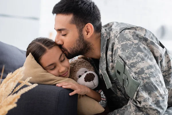 Man in military uniform kissing child with teddy bear on couch — Stock Photo