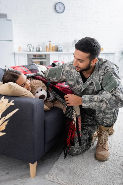 Man in camouflage uniform putting blanket on child with teddy bear on couch — Stock Photo