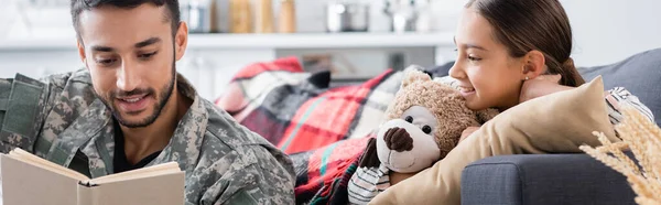 Smiling man in military uniform reading book near child with teddy bear on couch, banner — Stock Photo