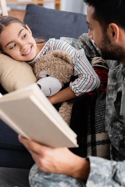 Enfant heureux avec un ours en peluche regardant le père flou en uniforme militaire tenant le livre à la maison — Photo de stock