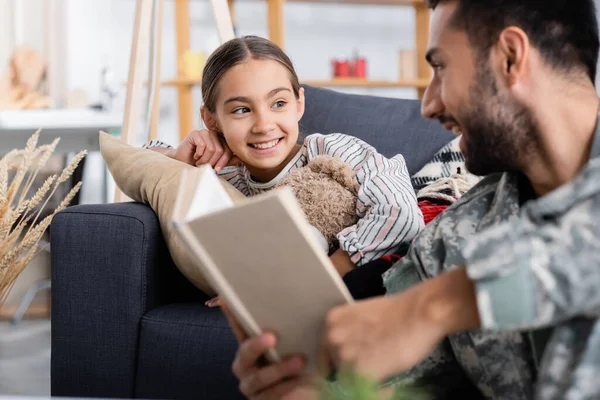 Smiling kid with teddy bear looking at blurred father in military uniform holding book at home — Stock Photo