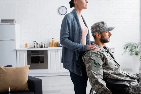 Donna in piedi vicino marito in uniforme militare e sedia a rotelle a casa — Foto stock