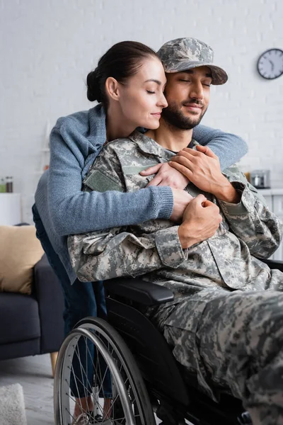 Mujer abrazando marido en uniforme de camuflaje y silla de ruedas en casa - foto de stock