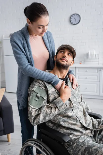 Mujer mirando al marido en uniforme militar cogido de la mano del marido en uniforme militar y silla de ruedas en casa - foto de stock