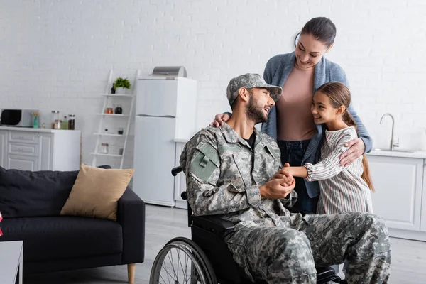 Enfant souriant tenant la main de papa en uniforme militaire en fauteuil roulant près de la mère à la maison — Photo de stock