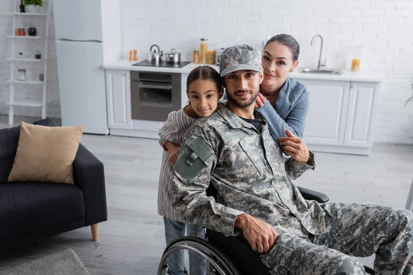 Homme en uniforme militaire et fauteuil roulant regardant la caméra près de la famille à la maison — Photo de stock