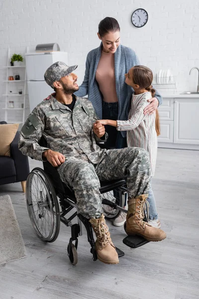 Fröhliche Frau und Kind stehen neben Vater in Militäruniform und Rollstuhl zu Hause — Stockfoto