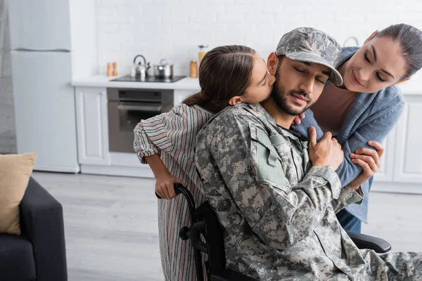 Niño besando padre en camuflaje uniforme y silla de ruedas cerca de mamá en casa - foto de stock